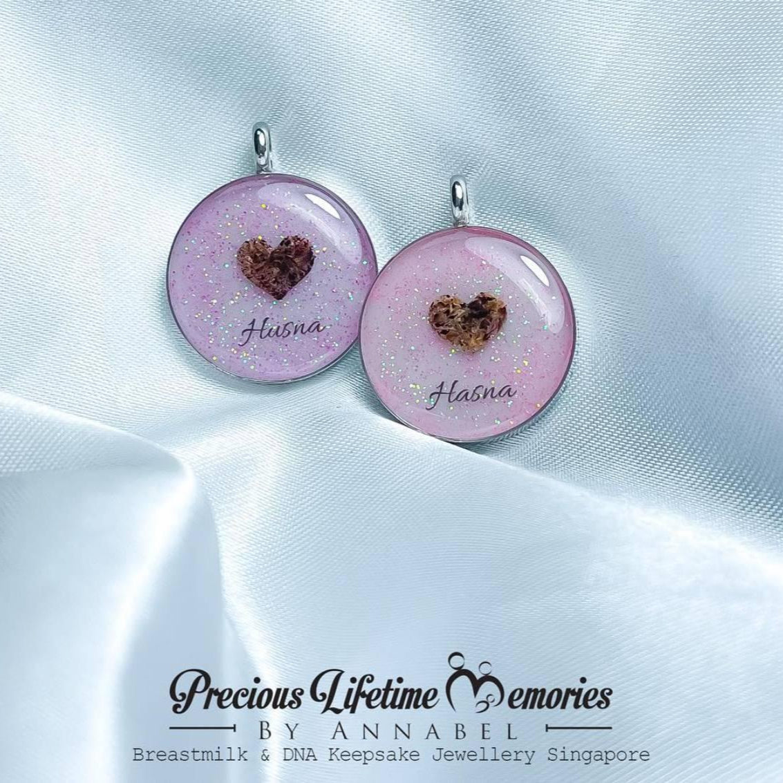 Pair of pink heart-shaped earrings with engraved names on a white satin background, branded 'Breastmilk Jewellery Singapore'.