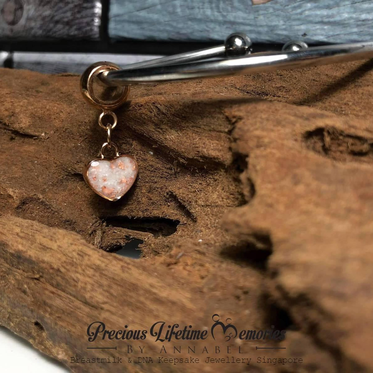 Bangle with a pets ashes heart-shaped charm on a wooden surface, featuring 'Precious Lifetime Memories by Annabel' branding.