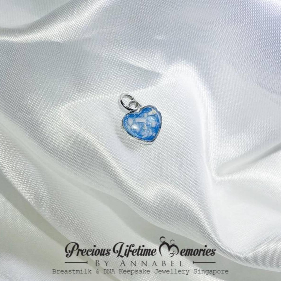 Heart-shaped blue pendant on white satin fabric with 'Precious Lifetime Memories by Annabel' branding.