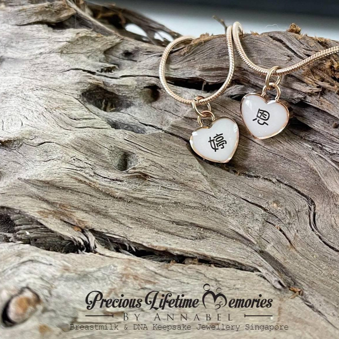 Two heart-shaped pendants on gold necklaces with engraved text, placed on driftwood.