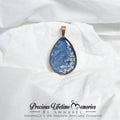 Teardrop-shaped blue pendant with gold edge  with pets fur and ashes keepsake jewellery on a white fabric background, branded 'Precious Lifetime Memories by Annabel'.