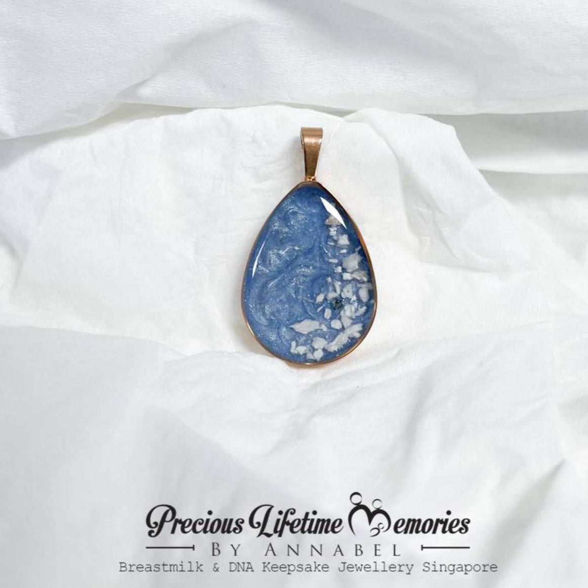 Teardrop-shaped blue pendant with gold edge  with pets fur and ashes keepsake jewellery on a white fabric background, branded 'Precious Lifetime Memories by Annabel'.