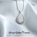 Teardrop-shaped pendant with pink and white glitter on a white satin background, featuring 'Precious Lifetime Memories by Annabel' branding.