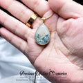 Teardrop-shaped pendant with blue and gold elements held in a hand, brand 'Precious Lifetime Memories' visible.
