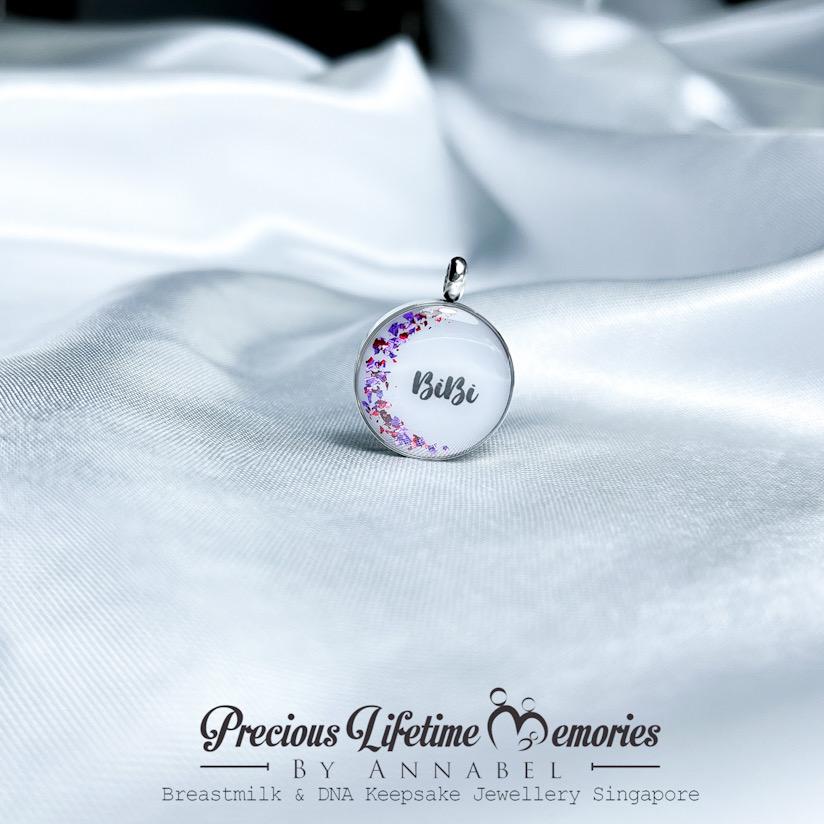 Silver pendant with floral design and 'Bibi' engraving on white fabric, branded 'Precious Lifetime Memories by Annabel'.