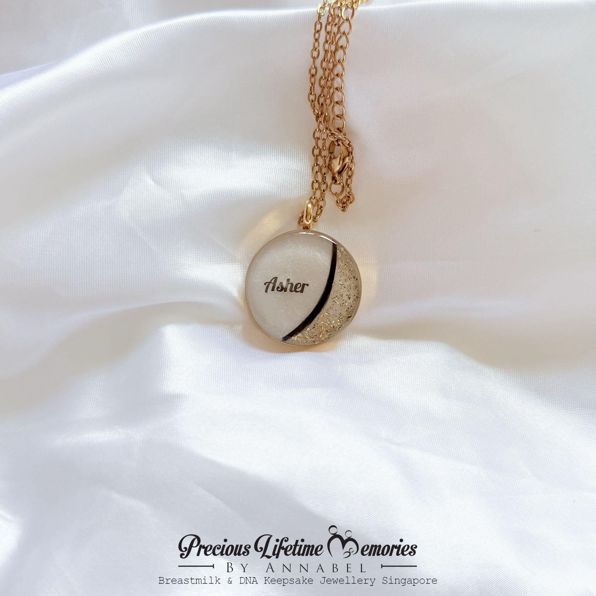Heart-shaped gold pendant with 'Asher' engraved on a white fabric background, branded 'Precious Lifetime Memories by Annabel'. breast milk with hair curl